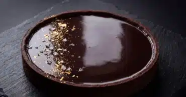 Une tarte au chocolat noir entière avec une ganache lisse et brillante comme un miroir, saupoudrée de quelques grains de fleur de sel, posée sur une ardoise.