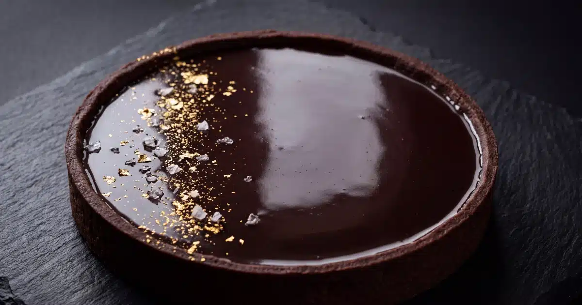 Une tarte au chocolat noir entière avec une ganache lisse et brillante comme un miroir, saupoudrée de quelques grains de fleur de sel, posée sur une ardoise.